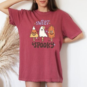 May include: A maroon-colored t-shirt with a retro Halloween design. The graphic features candy corn, ghost, and pumpkin popsicle illustrations with the text "Sweet & Spooky" in a playful font. The shirt is a comfortable, casual style.