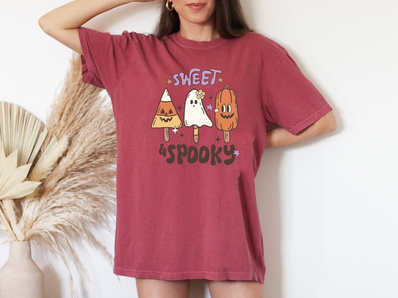 May include: A maroon-colored t-shirt with a retro Halloween design. The graphic features candy corn, ghost, and pumpkin popsicle illustrations with the text "Sweet & Spooky" in a playful font. The shirt is a comfortable, casual style.