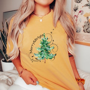 May include: A mustard yellow short-sleeve t-shirt featuring a watercolor-style green Christmas tree graphic with colorful ornaments. The text Merry & Bright is printed in black script above the tree, surrounded by decorative black swirling lines.