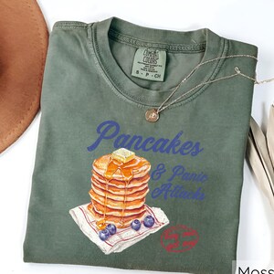 Op de afbeelding: Groen T-shirt met een afbeelding van een stapel pannenkoeken met siroop en boter, en de tekst "Pancakes & Panic Attacks".