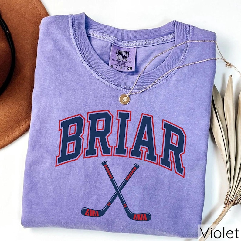 May include: A violet short-sleeved t-shirt with a crew neckline featuring the text BRIAR in dark blue with a red outline, positioned above a graphic of two crossed hockey sticks in dark blue with red accents. The shirt has a Comfort Colors label.