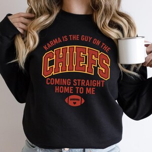 May include: Black sweatshirt with the text "Karma is the guy on the Chiefs, Coming straight home to me" in red and gold lettering. A football graphic with a heart is below the text. A white mug is held in the image.