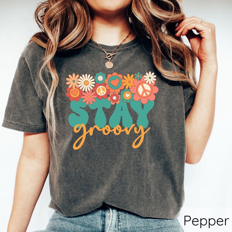 May include: A dark gray t-shirt with a vintage-inspired graphic design featuring a peace sign, hearts, and flowers. The text "Stay groovy" is printed in orange and teal.