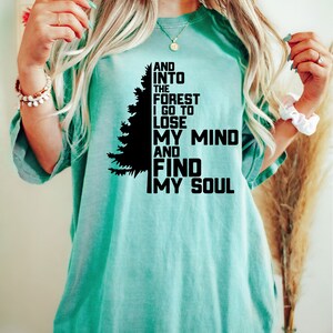 K&ouml;nnte beinhalten: Ein t&uuml;rkisfarbenes T-Shirt mit einer schwarzen Silhouette eines Kiefernbaums und dem Text "And into the forest I go to lose my mind and find my soul."