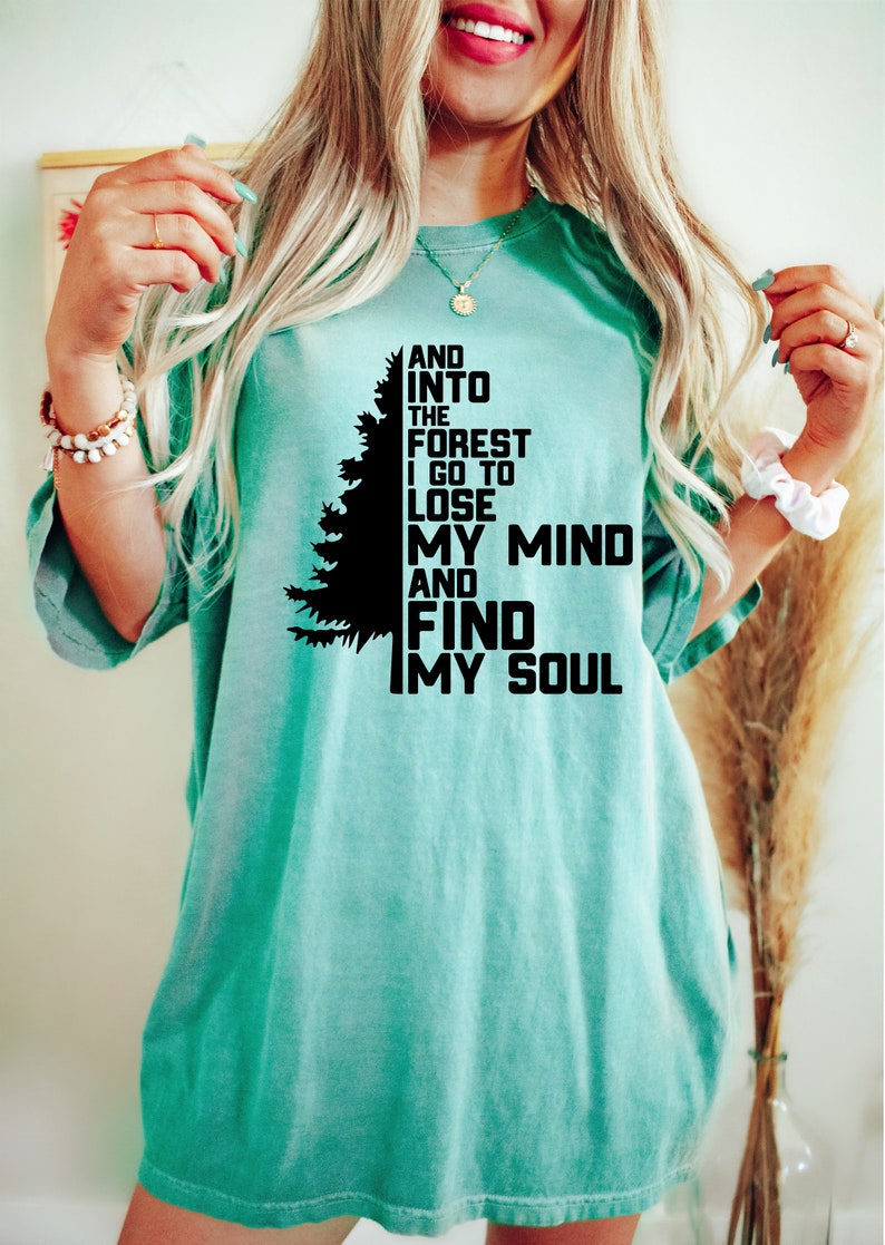K&ouml;nnte beinhalten: Ein t&uuml;rkisfarbenes T-Shirt mit einer schwarzen Silhouette eines Kiefernbaums und dem Text "And into the forest I go to lose my mind and find my soul."