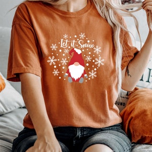 May include: A burnt orange short-sleeve t-shirt featuring a white graphic print of a gnome wearing a red hat, surrounded by white snowflakes and the text Let it Snow. The shirt has a crew neckline and a relaxed fit, paired with dark blue jeans.