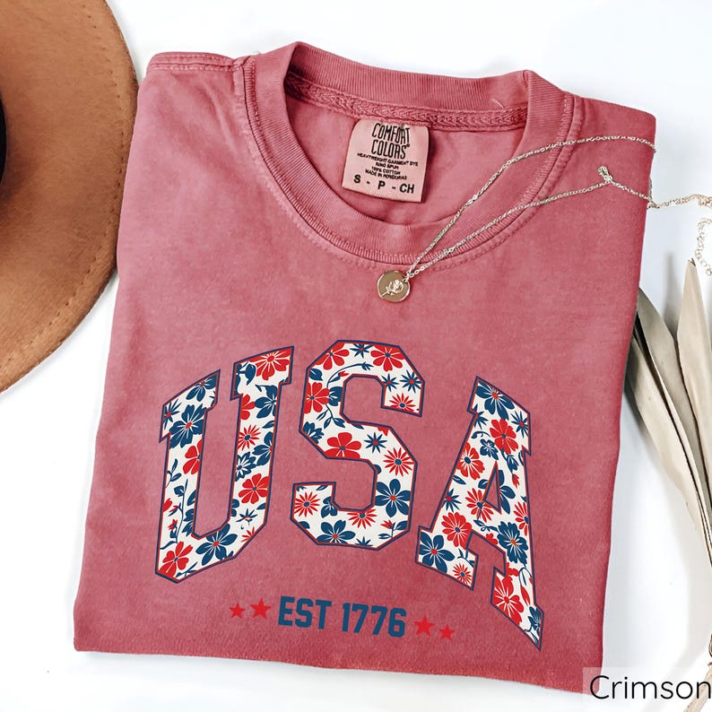 May include: Crimson-colored t-shirt with the word "USA" in a floral pattern. The shirt also features the text "EST 1776" and a small tag that reads "Comfort Colors". A silver necklace with a pendant is draped across the shirt.