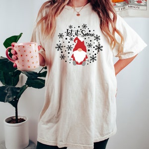 May include: An off-white short-sleeve t-shirt featuring a graphic of a gnome wearing a red hat and white beard, surrounded by black snowflakes and dots. The text "Let it Snow" is printed in black script above the gnome. The shirt is loose-fitting.