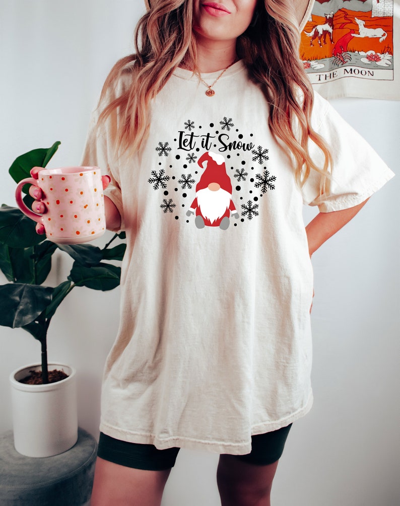 May include: An off-white short-sleeve t-shirt featuring a graphic of a gnome wearing a red hat and white beard, surrounded by black snowflakes and dots. The text "Let it Snow" is printed in black script above the gnome. The shirt is loose-fitting.