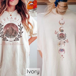 May include: Ivory t-shirt with a graphic design featuring a mountain range, moon phases, and butterflies. The text "Velaris City of Starlight" is printed on the front of the shirt.