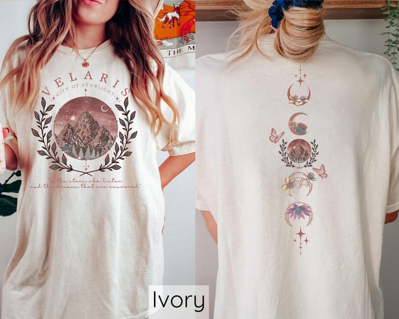 May include: Ivory t-shirt with a graphic design featuring a mountain range, moon phases, and butterflies. The text "Velaris City of Starlight" is printed on the front of the shirt.
