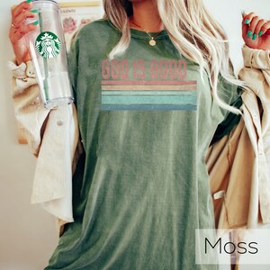 Puede incluir: Una camiseta verde con el texto "God is Good" en una fuente de estilo vintage con un dise&ntilde;o de rayas arco&iacute;ris. La camiseta est&aacute; hecha de un material suave y c&oacute;modo.