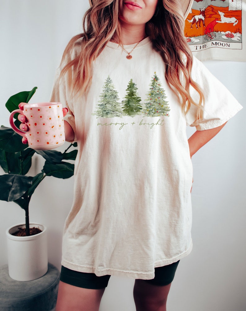 May include: Cream-colored t-shirt featuring a watercolor design of three Christmas trees. The trees are in shades of green, with the rightmost tree decorated with small ornaments. The words "merry + bright" are written in a script font below the trees.