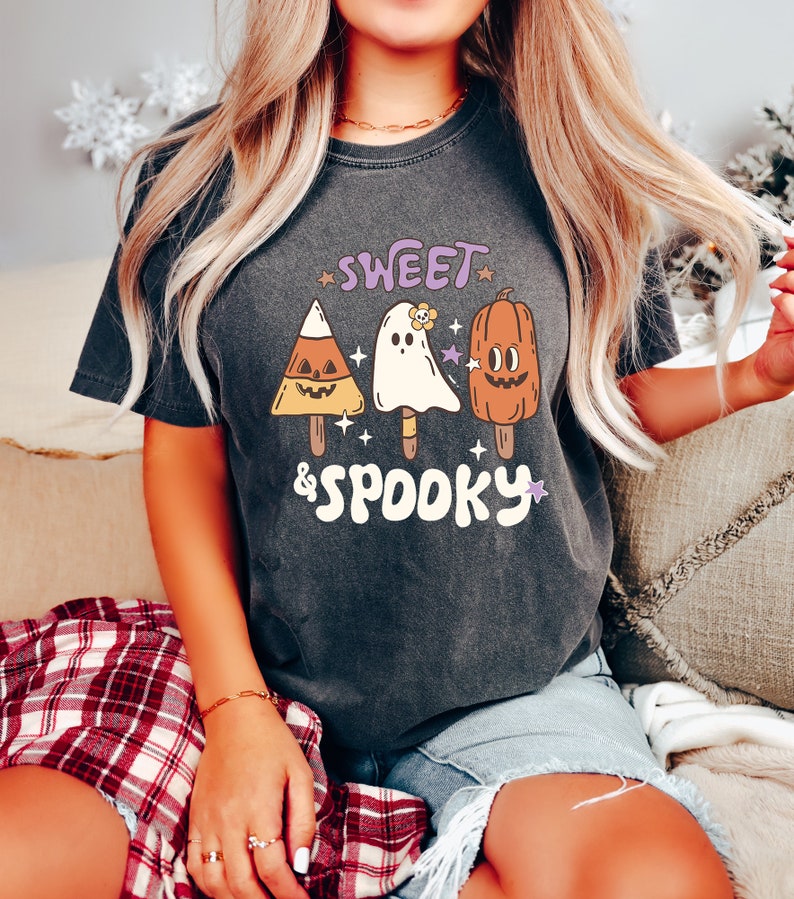 May include: Dark gray t-shirt with the words "Sweet & Spooky" and illustrations of Halloween-themed popsicles. The popsicles include a candy corn, a ghost, and a pumpkin. The shirt is a comfortable, casual style.