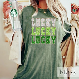 K&ouml;nnte beinhalten: &Uuml;bergr&ouml;&szlig;en-T-Shirt in Moosgr&uuml;n mit dem dreimal &uuml;bereinander angeordneten Text "LUCKY". Der Text ist in einer glitzernden Schriftart mit goldenen und gr&uuml;nen Akzenten gehalten.