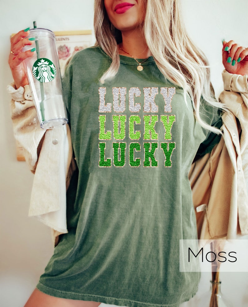 K&ouml;nnte beinhalten: &Uuml;bergr&ouml;&szlig;en-T-Shirt in Moosgr&uuml;n mit dem dreimal &uuml;bereinander angeordneten Text "LUCKY". Der Text ist in einer glitzernden Schriftart mit goldenen und gr&uuml;nen Akzenten gehalten.