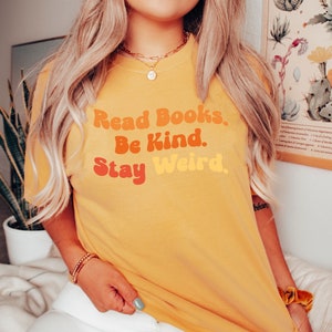 K&ouml;nnte beinhalten: Ein gelbes T-Shirt mit dem Text "Read Books. Be Kind. Stay Weird." in einer Retro-Schriftart mit orange, rot und gelben Farben.