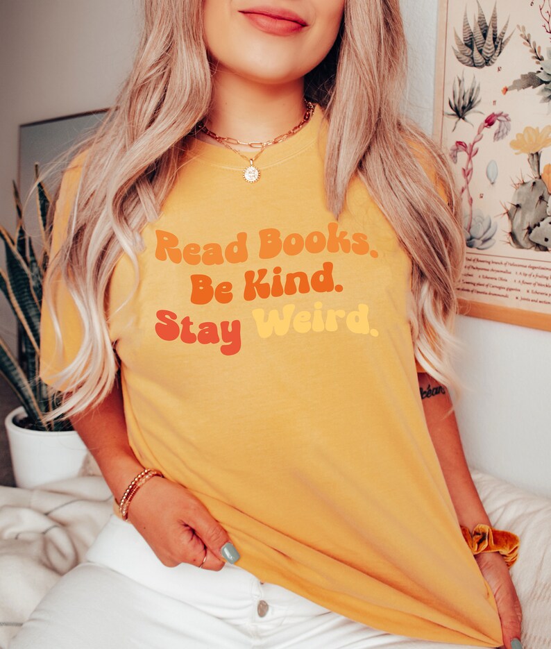 K&ouml;nnte beinhalten: Ein gelbes T-Shirt mit dem Text "Read Books. Be Kind. Stay Weird." in einer Retro-Schriftart mit orange, rot und gelben Farben.