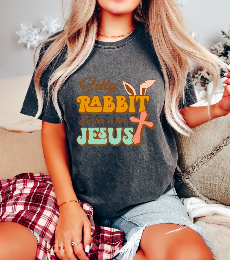 May include: A dark grey t-shirt with a retro-style graphic that says "Silly Rabbit Easter is for Jesus" with a pink bunny ear, orange bunny ear, and a pink cross.