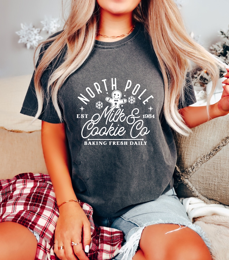 May include: Dark gray t-shirt with a white graphic that reads "North Pole Milk & Cookie Co. Baking Fresh Daily." The design includes a gingerbread man, snowflakes, and the establishment year, 1984. The shirt is short-sleeved and has a relaxed fit.