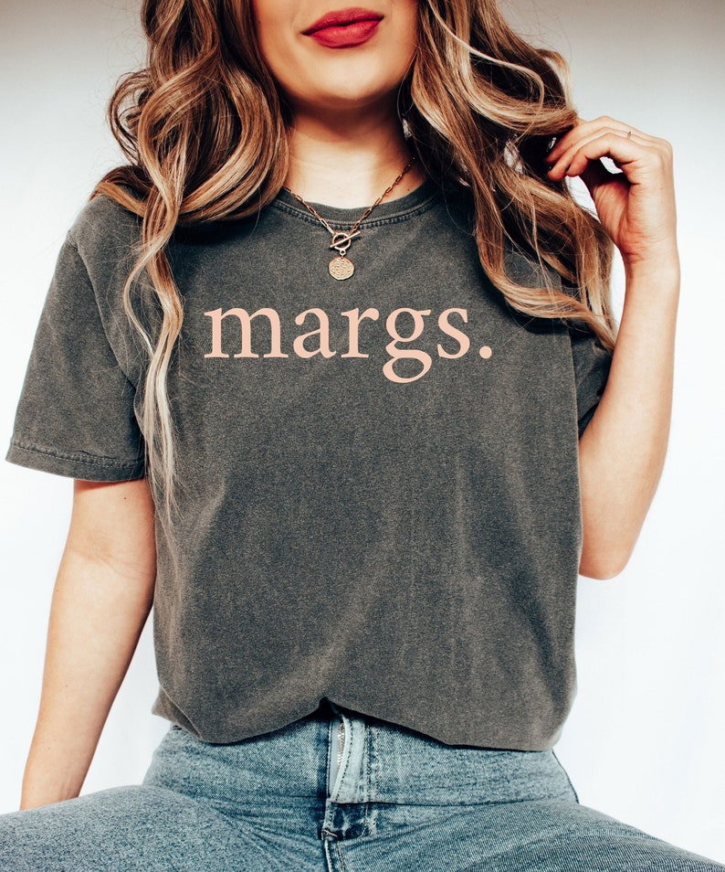 Puede incluir: Una camiseta gris oscura con la palabra "margs." impresa en rosa claro. La camiseta la lleva puesta una persona con el pelo largo y rubio y un collar de oro.