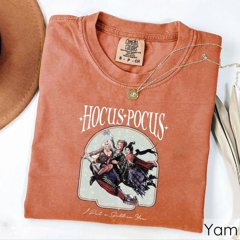Puede incluir: Una camiseta de color terracota con las palabras "Hocus Pocus" y un gr&aacute;fico de tres brujas. La camiseta tambi&eacute;n presenta la frase "I Put a Spell on You". Un collar de oro con un colgante est&aacute; drapeado sobre la camiseta.