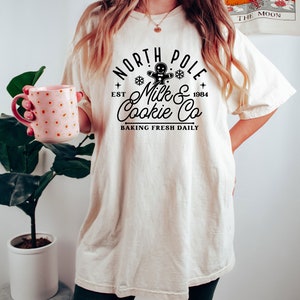 May include: Cream-colored t-shirt with a black graphic that reads "North Pole Milk & Cookie Co. Baking Fresh Daily." The design includes a gingerbread man, snowflakes, and the year 1984. The shirt is worn with dark green shorts.