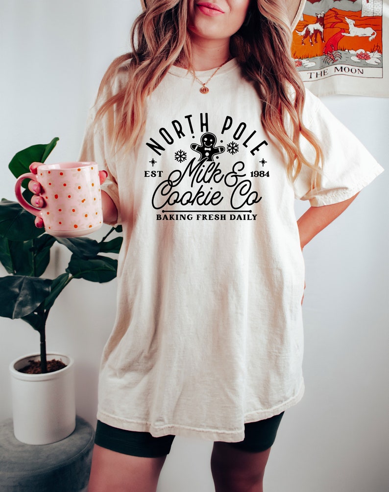 May include: Cream-colored t-shirt with a black graphic that reads "North Pole Milk & Cookie Co. Baking Fresh Daily." The design includes a gingerbread man, snowflakes, and the year 1984. The shirt is worn with dark green shorts.