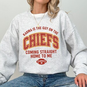 May include: A light gray sweatshirt with the text "Karma is the Guy on the Chiefs Coming Straight Home to Me" in red and gold lettering. A football graphic is below the text.