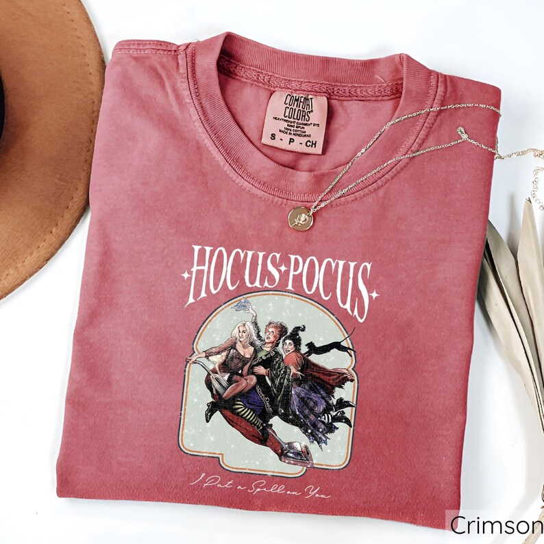 Puede incluir: Camiseta de color carmes&iacute; con las palabras "Hocus Pocus" y un gr&aacute;fico de tres brujas en una escoba. La camiseta tambi&eacute;n tiene la frase "I Put a Spell on You". Un collar dorado est&aacute; drapeado sobre la camiseta.