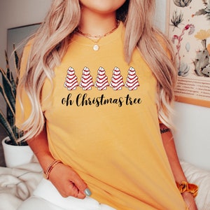 May include: Mustard yellow t-shirt featuring a graphic of five Christmas trees with red and white striped icing. The text "oh Christmas tree" is printed below the trees in black script. The shirt is a casual, festive apparel item.