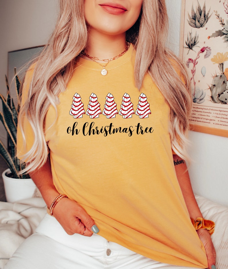 May include: Mustard yellow t-shirt featuring a graphic of five Christmas trees with red and white striped icing. The text "oh Christmas tree" is printed below the trees in black script. The shirt is a casual, festive apparel item.