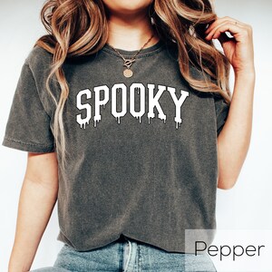 May include: A dark gray t-shirt with the word "SPOOKY" in white, dripping font. The shirt is a casual style, perfect for Halloween or spooky season. The model is wearing blue jeans.