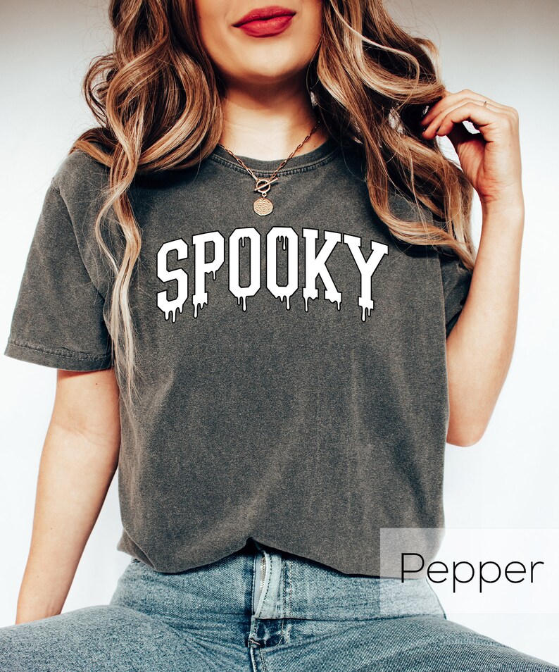 May include: A dark gray t-shirt with the word "SPOOKY" in white, dripping font. The shirt is a casual style, perfect for Halloween or spooky season. The model is wearing blue jeans.