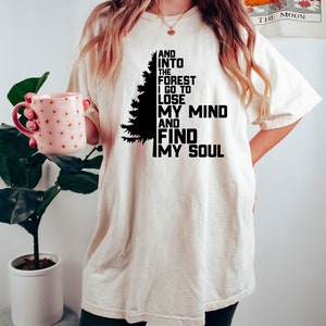 K&ouml;nnte beinhalten: Ein wei&szlig;es T-Shirt mit einer schwarzen Silhouette eines Kiefernbaums und dem Text "AND INTO THE FOREST I GO TO LOSE MY MIND AND FIND MY SOUL".