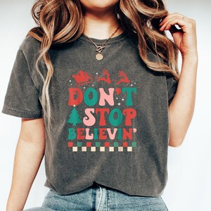 May include: A charcoal gray short-sleeve t-shirt featuring a red, pink, and green graphic print. The design includes a silhouette of Santa in a sleigh with reindeer, a Christmas tree, and the text "DON'T STOP BELIEVIN'" above a checkered pattern.