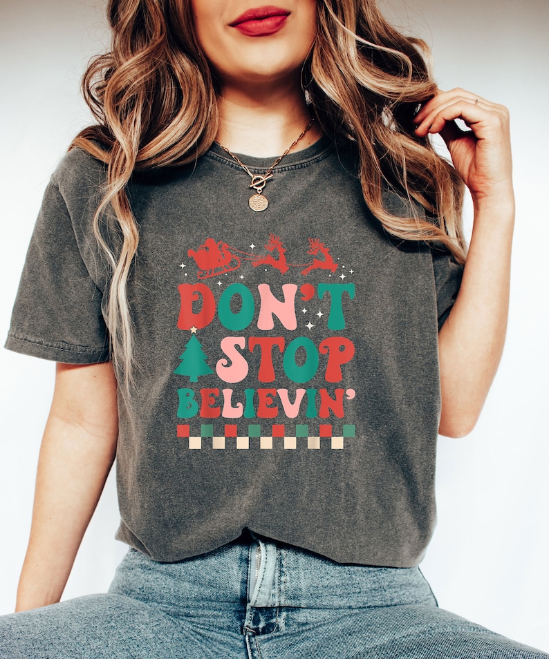 May include: A charcoal gray short-sleeve t-shirt featuring a red, pink, and green graphic print. The design includes a silhouette of Santa in a sleigh with reindeer, a Christmas tree, and the text "DON'T STOP BELIEVIN'" above a checkered pattern.