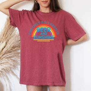 May include: A maroon t-shirt with the text "THE ONLY B.S. I NEED IS BEER AND SUNSHINE" in a rainbow design. The shirt is a relaxed fit and has short sleeves.