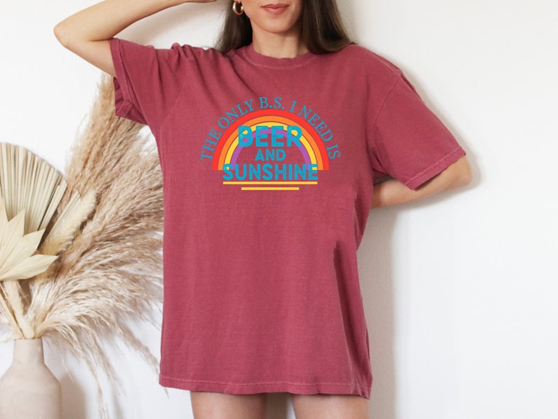 May include: A maroon t-shirt with the text "THE ONLY B.S. I NEED IS BEER AND SUNSHINE" in a rainbow design. The shirt is a relaxed fit and has short sleeves.