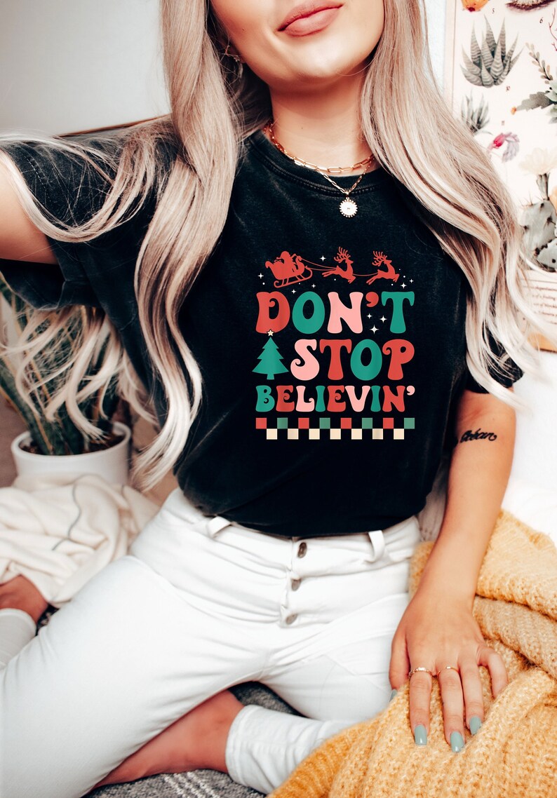 May include: A black short-sleeve t-shirt featuring a graphic print with the text "DON'T STOP BELIEVIN'" in red, pink, and green. The design includes a silhouette of Santa in a sleigh with reindeer, a small Christmas tree, and a checkered border.