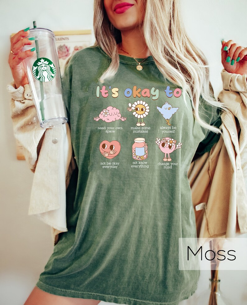 K&ouml;nnte beinhalten: Gr&uuml;nes T-Shirt mit dem Text "It's okay to" und Illustrationen eines Gehirns, einer Sonne, eines Vogels, eines Herzens, eines Glases und eines Gesichts. Der Text unter jeder Illustration lautet: "need your own space", "make some mistakes", "always be yourself", "not be okay everyday", "not know everything", und "change your mind".