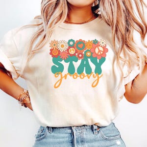 May include: A white t-shirt with a graphic design featuring a cluster of colorful flowers, peace signs, and hearts. The text "STAY groovy" is written in a retro font in teal and orange.