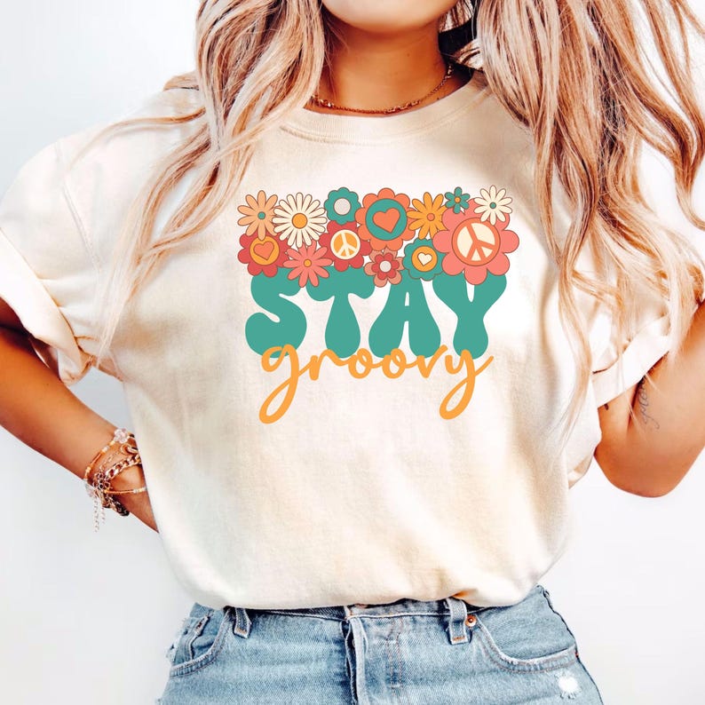 May include: A white t-shirt with a graphic design featuring a cluster of colorful flowers, peace signs, and hearts. The text "STAY groovy" is written in a retro font in teal and orange.