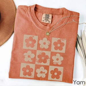 May include: A terracotta-coloured t-shirt with a vintage-style design of nine white daisy flowers in a grid pattern. The shirt has a tag that reads "Comfort Colors" and "S - P - CH".