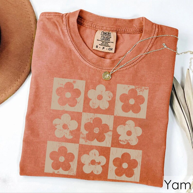 May include: A terracotta-coloured t-shirt with a vintage-style design of nine white daisy flowers in a grid pattern. The shirt has a tag that reads "Comfort Colors" and "S - P - CH".