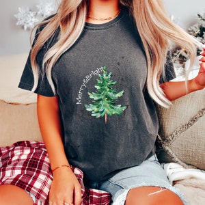 May include: A charcoal gray short-sleeve t-shirt featuring a watercolor-style green Christmas tree graphic with colorful ornaments and the text Merry & Bright printed in white script lettering on the front of the garment.