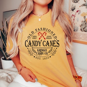 Pu&ograve; includere: T-shirt giallo senape con una grafica circolare che recita "Old Fashioned Candy Canes" con illustrazioni di bastoncini di zucchero. Il testo include anche "Kringle Candy Co. Est. 1822".