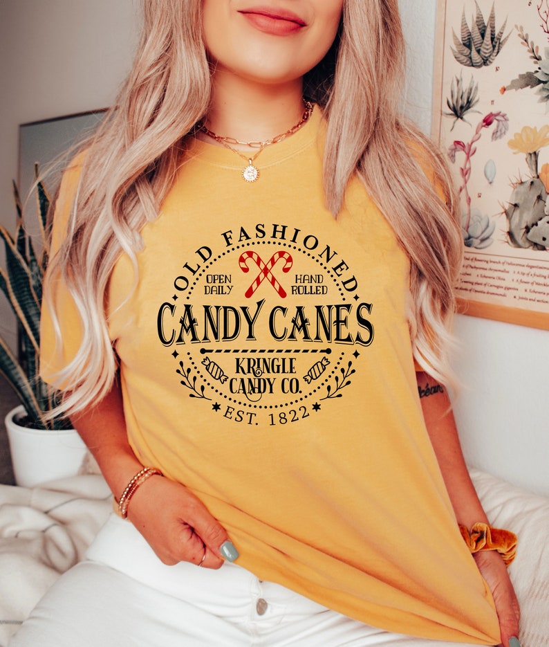 Pu&ograve; includere: T-shirt giallo senape con una grafica circolare che recita "Old Fashioned Candy Canes" con illustrazioni di bastoncini di zucchero. Il testo include anche "Kringle Candy Co. Est. 1822".