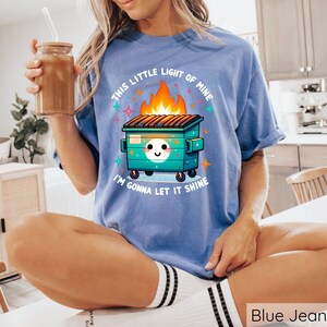 May include: A blue t-shirt with a graphic of a green dumpster with a smiling face and flames coming out of the top. The text "This little light of mine I'm gonna let it shine" is printed on the shirt.