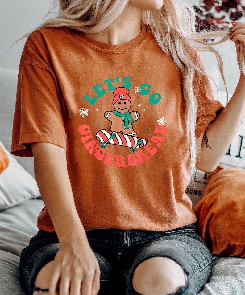 May include: Burnt orange t-shirt with a graphic that says "Let's Go Gingerbread." The graphic features a gingerbread man on a skateboard, surrounded by snowflakes. The shirt is short-sleeved and has a relaxed fit.